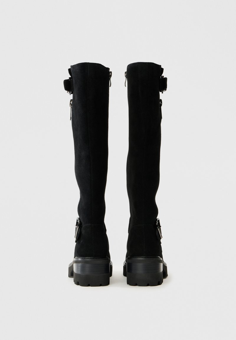 Botas altas de ante negro con tacón grueso, que cuentan con cremalleras y hebillas decorativas en los laterales. Textura suave, diseño elegante.