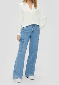 Blusa bianca leggera con scollatura a V e maniche lunghe, abbinata a jeans cargo azzurri morbidi con tasche laterali. Sneakers bianche.