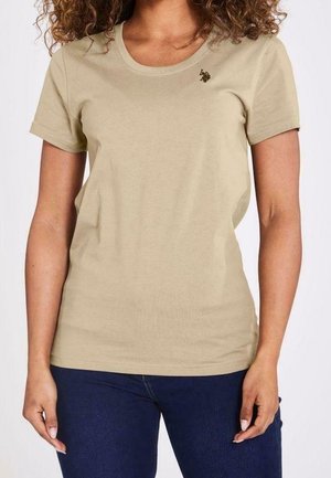 Mujer con camiseta beige de manga corta con un pequeño logo negro bordado en el lado izquierdo del pecho y jeans azul oscuro.