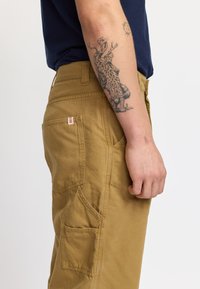 Pantaloni cargo beige con molte tasche, cuciture a punto, vestibilità rilassata e tessuto di cotone robusto. Presenta un piccolo etichetta con il marchio vicino alla vita.
