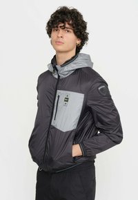 Giacca leggera con cappuccio nera e grigia, presenta una chiusura frontale con zip, polsini elastici e una tasca sul petto con dettagli metallici.