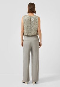 Kobieta w bezrękawnikowej srebrnej bluzce z cekinami i jasnoszarych szerokich spodniach, stojąca tyłem na jednolitym tle.