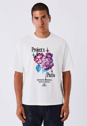Camiseta de algodón blanca con un gráfico floral en rosa y púrpura, con texto negro que dice "Project x Paris" y "aventura comunitaria."