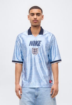 Tânăr care poartă tricou de fotbal Nike al Angliei, albastru deschis, cu guler închis și blugi deschiși, stând pe fundal alb.
