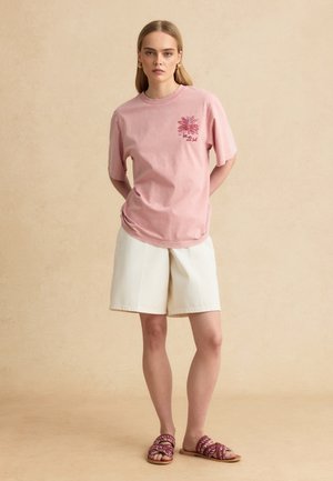 Femme debout, les mains dans le dos, portant un t-shirt graphique rose, un short blanc aux genoux, et des sandales à enfiler à motifs sur fond beige.
