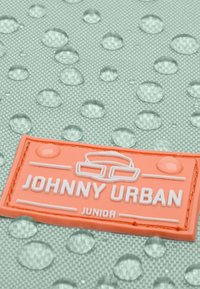 Zelená texturovaná látka s kapkami vody a oranžovým gumovým logem, na kterém je napsáno „JOHNNY URBAN JUNIOR.“