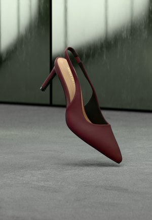 Bordeauxfarbener Lederslingback-Pumps mit spitz zulaufender Schuhspitze und schlankem Absatz, der auf einer grauen Oberfläche neben einem reflektierenden Fenster liegt.