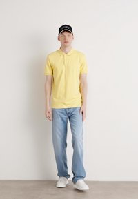 Gul polo skjorte med krage, uformelle blå jeans og hvite joggesko. En svart caps med "BOSS" logo fullfører antrekket.