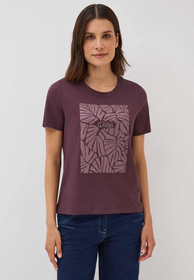 T-shirt en coton violet avec un col rond et des manches courtes. Présente un graphique carré de feuilles de palmier dans une teinte plus claire avec du texte en dessous.