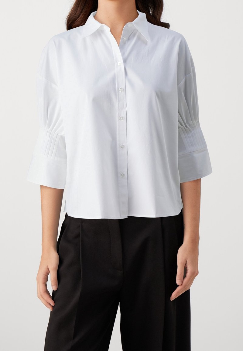 Camisa blanca con un cuello estructurado, botones en la parte delantera y mangas tres cuartos con pliegues. La tela parece suave y ligera. Combinada con pantalones negros.