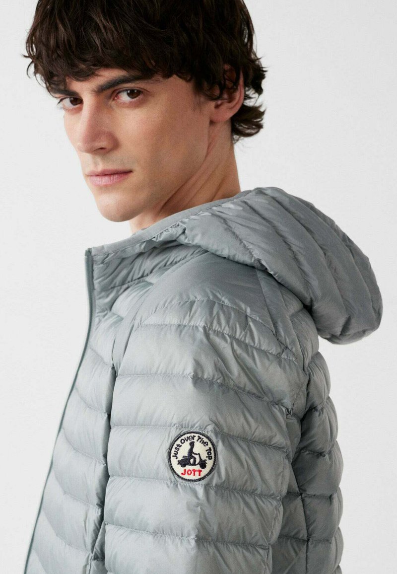 Joven con chaqueta acolchada gris claro con capucha, mostrando un parche redondo con un scooter y texto en la parte superior del brazo.