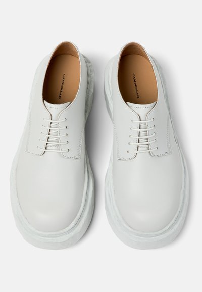 Pari valkoisia nahkaisia nauhallisia kenkiä, joissa paksut kuvioidut pohjat ja vaaleanruskeat sisäpohjat, joissa lukee "Camperlab", esitetty ylhäältä valkoisella taustalla.