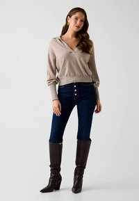 Pull-over beige en maille avec col en V et manches longues, associé à un jean skinny bleu foncé avec des boutons dorés. Bottes marron montant jusqu'aux genoux.