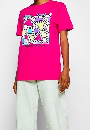 Jasnoróżowa koszulka z kolorowymi geometrycznymi wzorami w stylu lat 80. i napisem „BORN IN THE 80’S”, noszona z jasnymi spodniami.