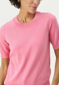 Pull à col rond à manches courtes en tissu rose doux. Texture lisse avec des bords côtelés au niveau du col et des manches. Présente une coupe décontractée.