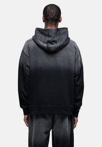 Felpa nera con gradiente, dal tessuto morbido, grande cappuccio e taglio ampio, con polsini e orlo a coste. Indossata con pantaloni della tuta scuri abbinati.