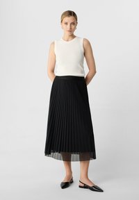 Zwarte plissee rok met een doorschijnende onderlaag, gecombineerd met een witte geribde tanktop. Zwarte puntige slingback schoenen maken de outfit compleet.