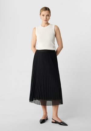 Gonna nera a pieghe con uno strato inferiore trasparente, abbinata a una canotta bianca a coste. Scarpe nere con punta a slingback completano l'outfit.