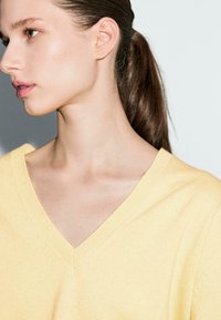 Gelber V-Ausschnitt-Pullover aus Strickmaterial, mit lockerer Passform und weicher Textur, vor einem hellen Hintergrund aufgenommen.
