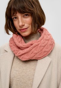 Écharpe infinity en laine corail tricotée avec un motif de câble texturé, portée sur un pull beige côtelé et un manteau de couleur claire.