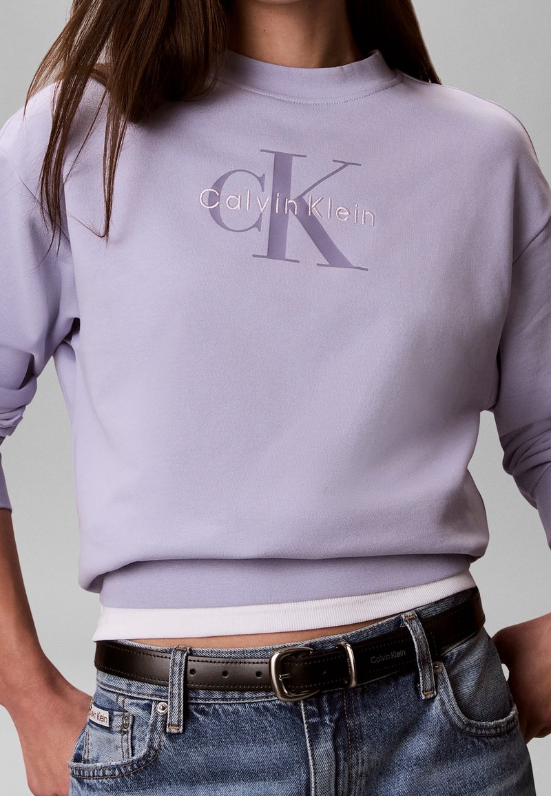 Personne portant un sweat-shirt lavande Calvin Klein avec le logo CK, associé à un jean bleu et une ceinture en cuir noire.