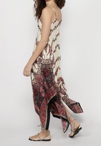 Femme marchant dans une robe maxi crème sans manches avec des motifs cachemire rouges et noirs, portant des sandales plates noires à lanières.