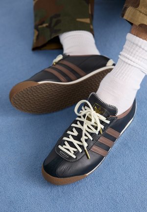 Czarne i brązowe trampki Adidas z białymi sznurówkami, noszone z białymi skarpetkami i spodniami moro na niebieskim dywanie.
