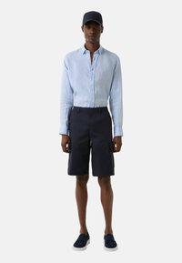 Lyseblå linned skjorte, marineblå shorts med cargo-lommer og marineblå kasket. Sættet har et afslappet, tætsiddende design og elegante slip-on sko.