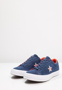 Zapatillas de ante azul marino con suelas de goma blanca, que presentan acentos rojos y un logo de estrella blanca en el lateral. Diseño de punta redondeada y con cordones.