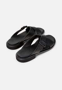 Black leather sandals with a flat sole, featuring a toe post design, adjustable buckle, and engraved logo on the footbed.