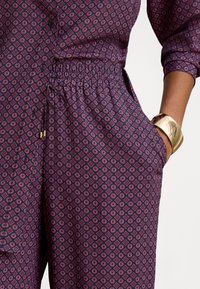 Midje og hofte til en person som har på seg lilla mønstrede bukser og skjorte, med hånden i lommen og et stort gullarmbånd på håndleddet.