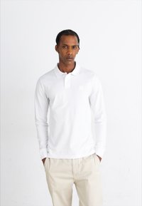 Tricou polo alb cu mânecă lungă, realizat din bumbac, având o garnitură cu trei butoane și un logo mic pe pieptul stâng. O pereche de pantaloni bej.