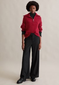 Roter, gerippter Strickpullover mit einem Reißverschlusskragen, getragen über einem schwarzen Rollkragenpullover. Kombiniert mit weiten schwarzen Hosen und klobigen schwarzen Schuhen.