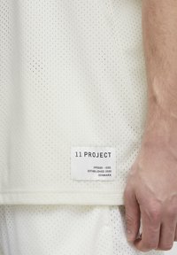 Hvit, pustende mesh-skjorte med små hull, med en etikett som inkluderer "11 PROJECT" og vaskeanvisninger på sømmen.