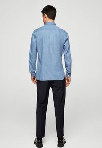 Hellblaues Jeanshemd mit langen Ärmeln, geknöpften Manschetten, klassischem Kragen und abgerundetem Saum; kombiniert mit dunkler Hose und schwarzen Schuhen.