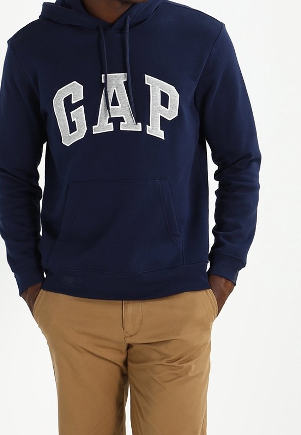 Personne portant un sweat à capuche bleu marine avec logo blanc GAP et pantalon beige, mains dans les poches, sur un fond clair uni.