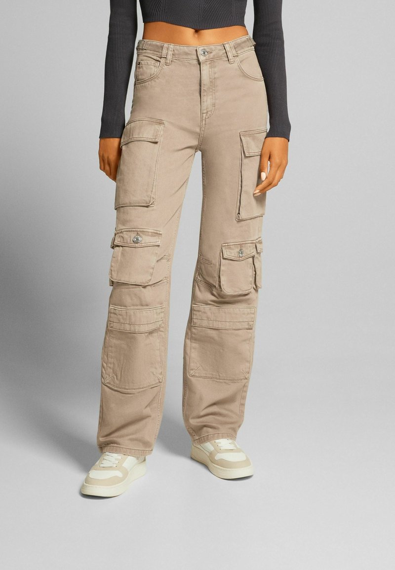 Pantalones cargo beige con grandes bolsillos laterales, fabricados de una tela con textura suave. Presenta un cierre de botón y un ajuste relajado.
