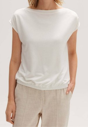 Valkoinen lyhythihainen paita, jossa on veneen kaula-aukko ja resorimainen helma, valmistettu pehmeästä kankaasta. Yhdistetty vaaleanbeigeihin raidallisiin housuihin, kädet taskuissa.