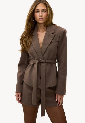 Femme portant un blazer marron ceinturé et un short assorti, avec des boucles d'oreilles anneaux, posant devant un fond blanc uni.