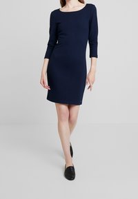 Robe côtelée bleu marine avec un col bateau et des manches trois-quarts, présentant une silhouette ajustée et une longueur au genou. Portée avec des ballerines noires.