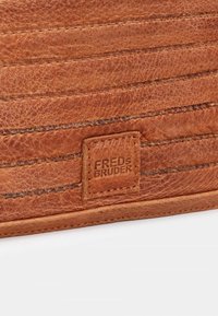 Portafoglio in pelle marrone caratterizzato da linee cucite verticalmente e un'etichetta quadrata impressa con "FREDS BRUDER" per il branding. Dettagli con superficie testurizzata.