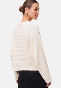 Femme avec les cheveux en chignon, portant un pull en maille crème texturé et une jupe noire, tournée légèrement sur le côté devant un fond uni.