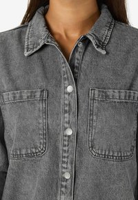 Hallhallane teksast särk, millel on krae, nööbid ja kaks rinna taskut. Omab nähtavat õmblustDetaili ja pehmet tekstuuri.