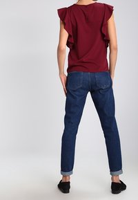 Bordeaux-top med flæser på ærmerne og knapper bagpå, parret med blå denim jeans, der har opfoldede manchetter og lommedesign. Sorte sko fuldender looket.