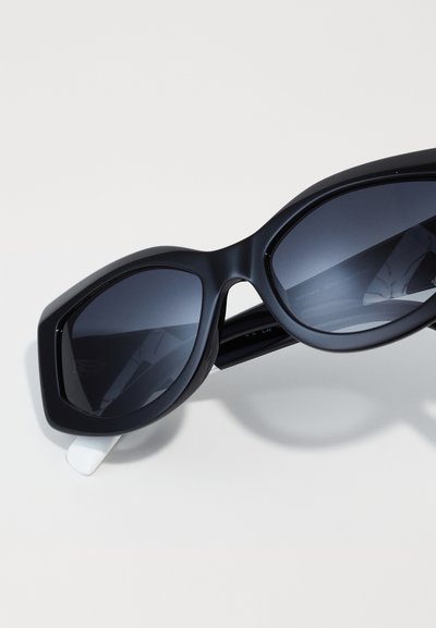 Black oversized sunglasses with dark lenses, thick, angular plastic frame, and a white accent on the temple. Smooth texture, geometric design.