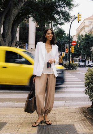 Kvinde i hvid blazer og beige bukser holder kaffe og taske, står ved fodgængerfelt med gul bil og trafiklys i baggrunden.