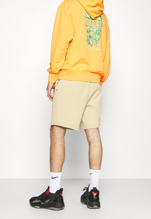 Person trägt einen orangefarbenen Hoodie mit einem Basketball-Logo auf dem Rücken, beige Shorts, weiße Nike-Socken und schwarze Sneaker und steht mit dem Rücken zu uns.