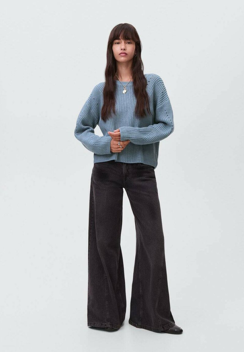 Přehozený světle modrý pletený svetr s vroubkovanou strukturou, v kombinaci s černými širokými kalhotami s vysokým pasem. Modelka stojí se složenýma rukama.