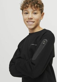 Svart sweatshirt med rund halsringning och långa ärmar, med en ficka med dragkedja på vänster arm och logotypdetalj ovanför fickan.