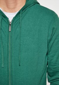 Grüner Zip-Hoodie mit strukturiertem Muster, verstellbarem Kapuzenzug und silbernem Reißverschluss.